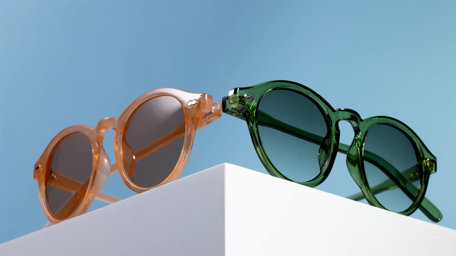 Two pairs of sunglasses, one orange and one green, on a white surface with a blue sky background.